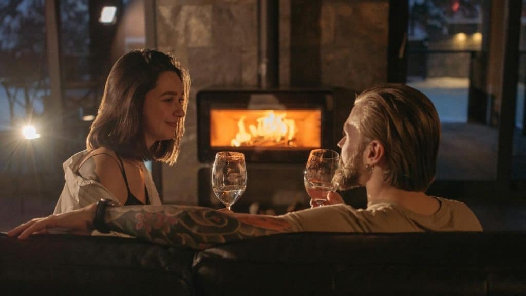 A man and woman sit on a sofa holding wine glasses near a glowing fireplace.
