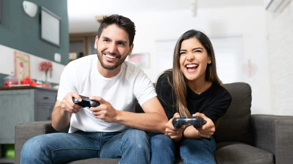 A laughing man and woman sit on a grey sofa holding black video game controllers.