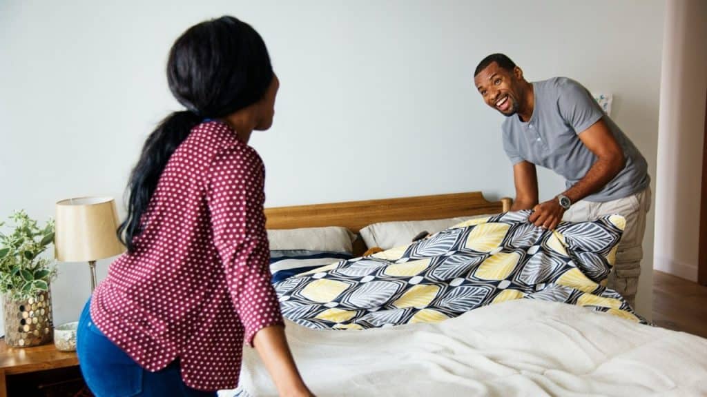 A smiling man and woman work together to spread patterned sheets across a wooden bed.