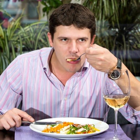 A man in a striped shirt sits at a restaurant table eating a meal with wine.