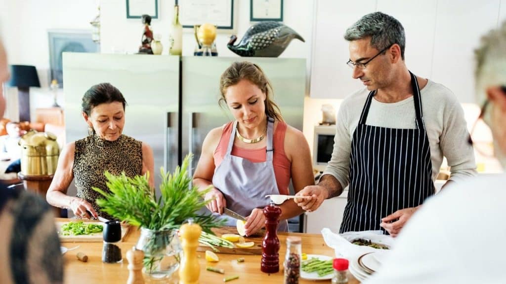 Three people prepare food at a wooden counter in a kitchen while wearing aprons.