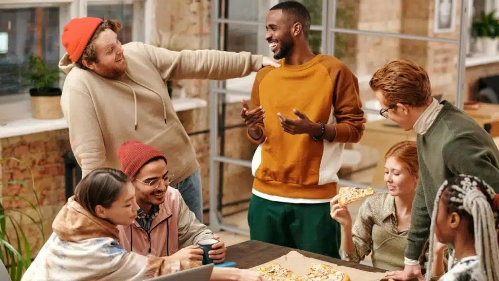 A group of friends laughs and eats pizza together around a wooden table indoors.