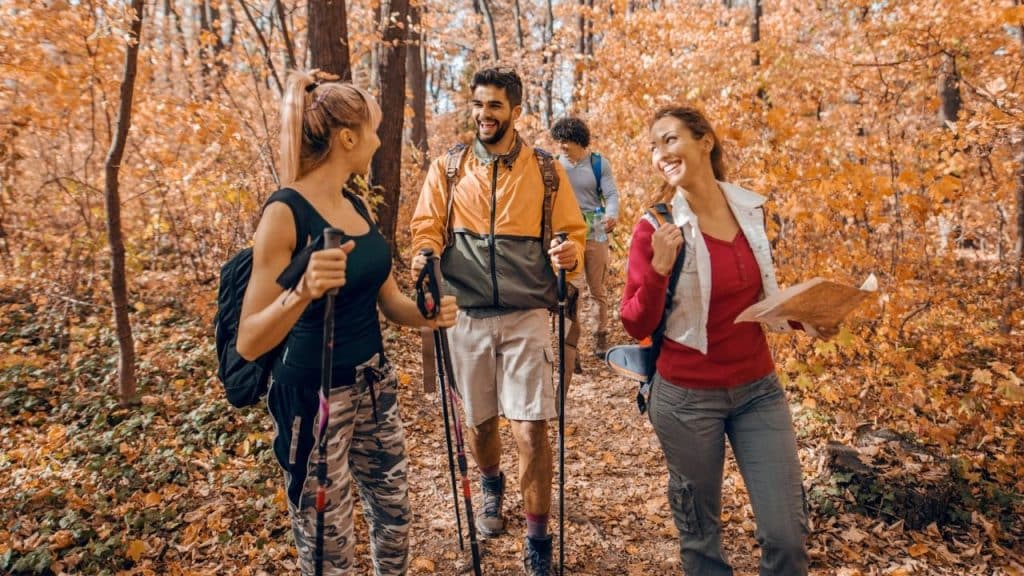 Three people with backpacks and walking sticks hike through a forest with orange autumn leaves.