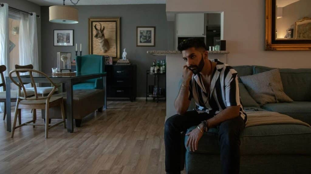 A man in a striped shirt sits on a sofa, resting his chin on his hand.