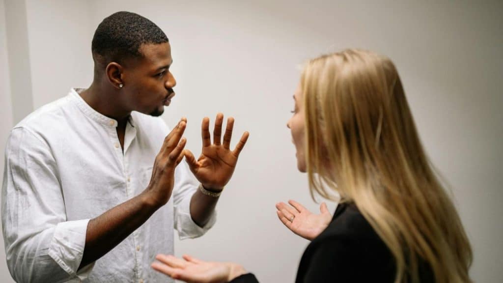 A man and woman stand facing each other, gesturing with their hands during a conversation.