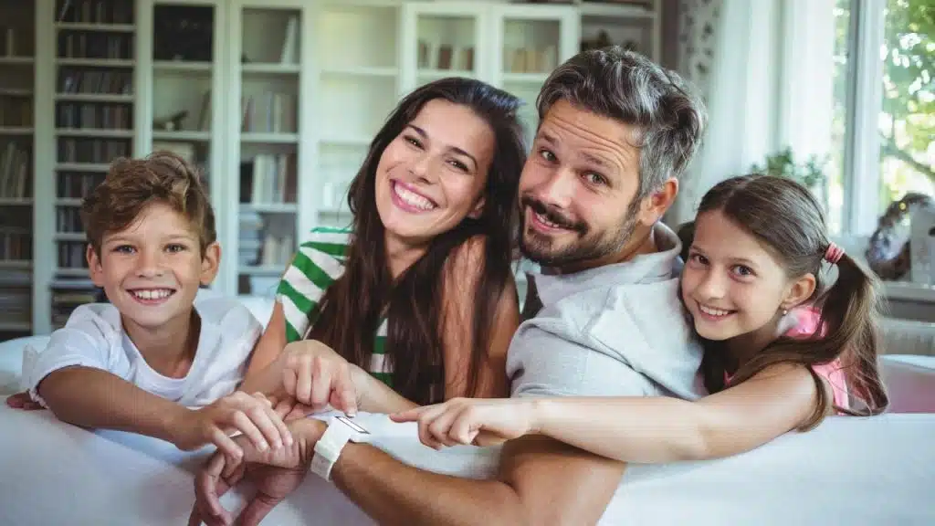 A smiling family of four leans over a white sofa, pointing at a white smartwatch.