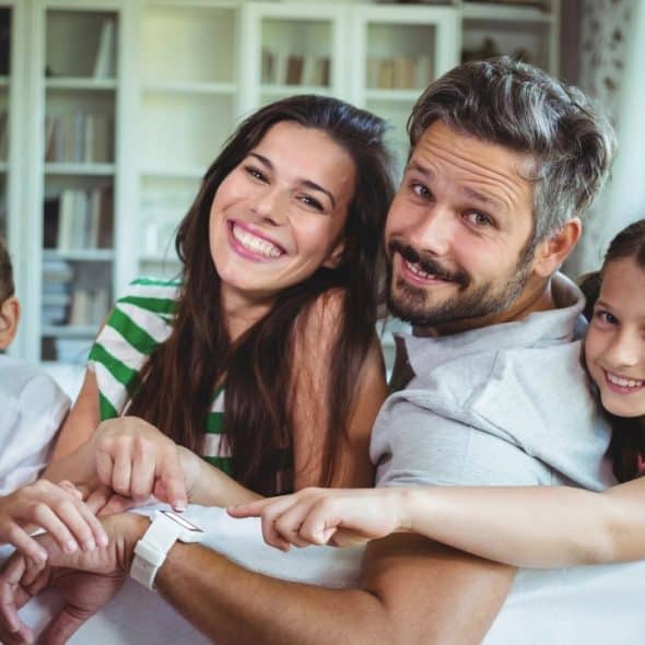 A smiling family of four leans over a white sofa, pointing at a white smartwatch.