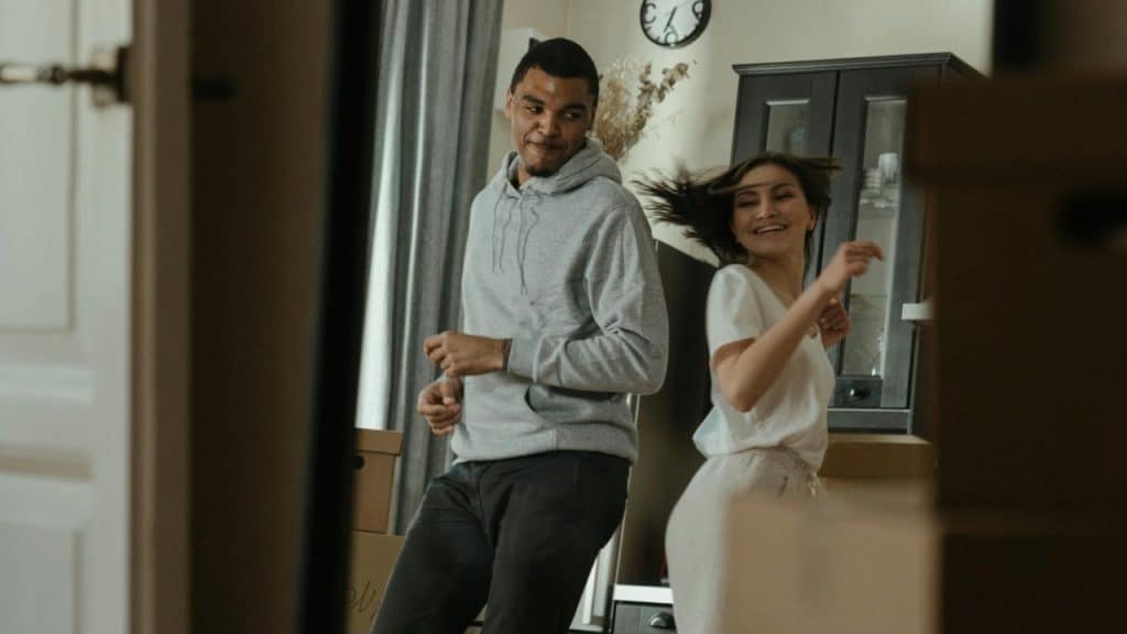 A man and woman dance in a living room filled with several cardboard boxes.