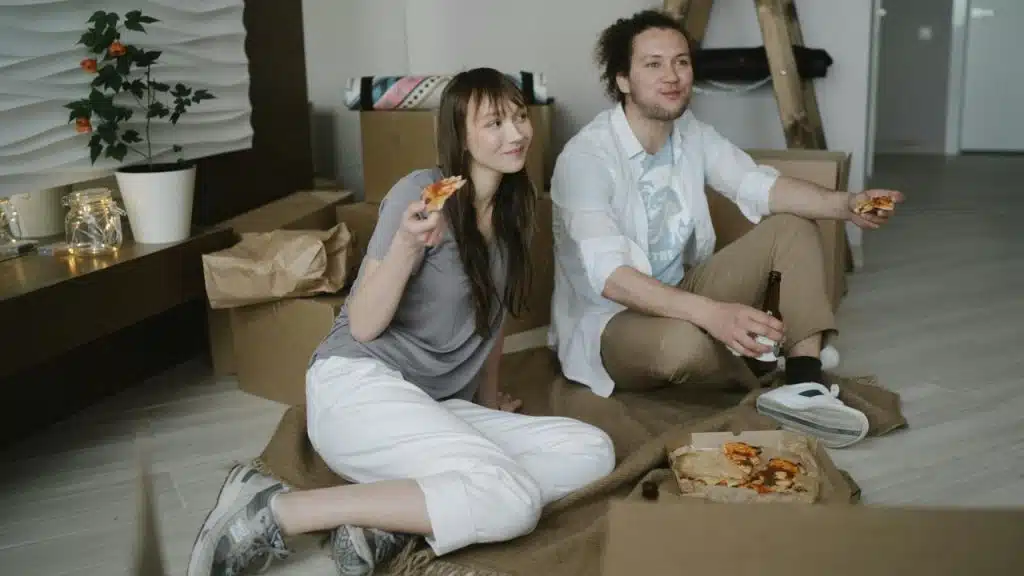 A man and woman sit on a floor blanket eating pizza near several cardboard boxes.
