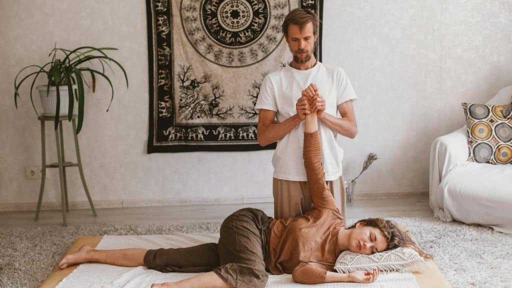 A man performs a hand and arm stretch on a woman lying on a floor mat.