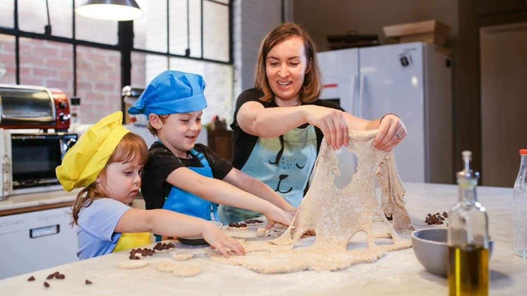 A woman and two children in chef hats prepare dough with chocolate chips on a counter.