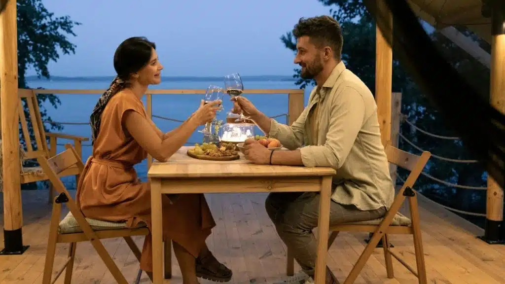 A man and woman sit at a wooden table outdoors, clinking wine glasses at dusk.