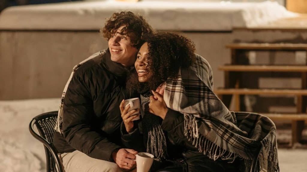 A man and woman huddle under a blanket outdoors at night, holding white mugs.