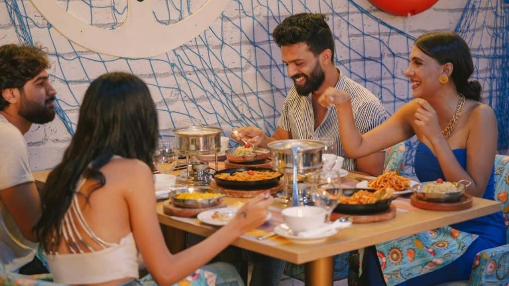 Two couples sit around a table eating various pasta dishes in a nautical-themed room.