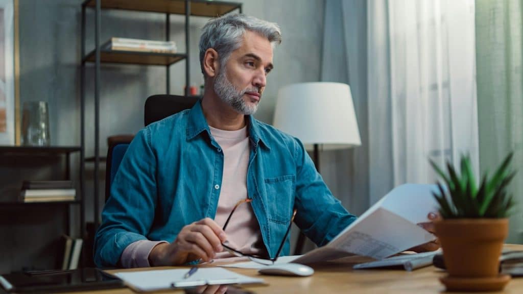 A middle-aged bearded man tending to financial documents and looking perturbed.