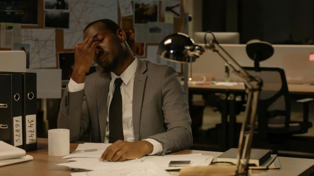 A man in a suit sits at a desk with papers, rubbing the bridge of his nose.