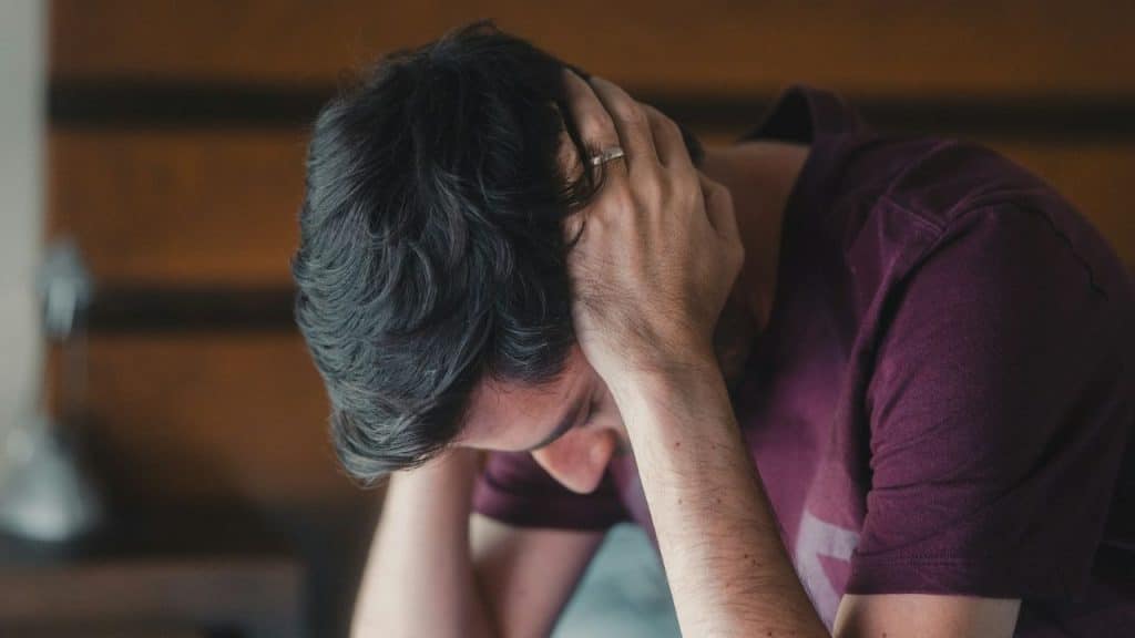 A man in a maroon shirt sits with his head down, holding it with both hands.