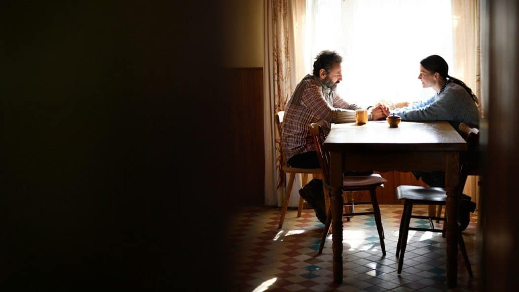 A man and woman sit facing each other at a wooden dining table, holding hands in a room brightly lit by sunlight.
