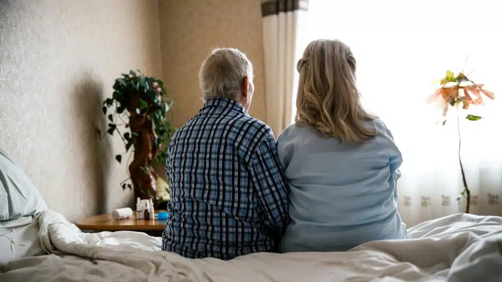 The back view of an older couple sitting together on the edge of a bed, looking toward a bright window.