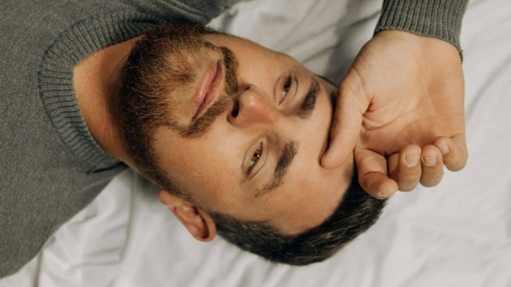 A bearded man in a gray sweater lies on white bedding, touching his forehead.