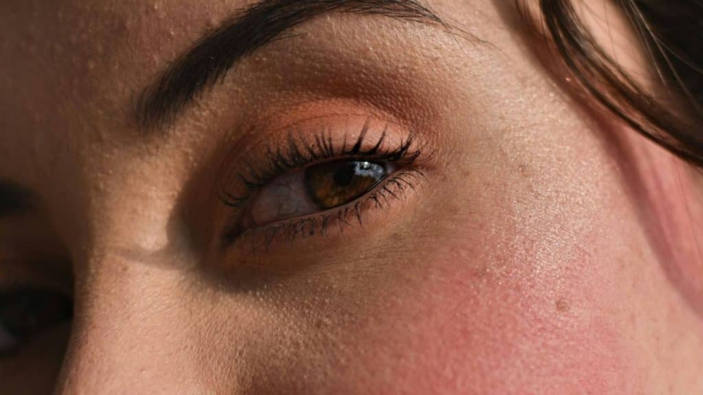 A close-up view of a person's eye and face featuring warm, neutral-toned makeup and sunlight.