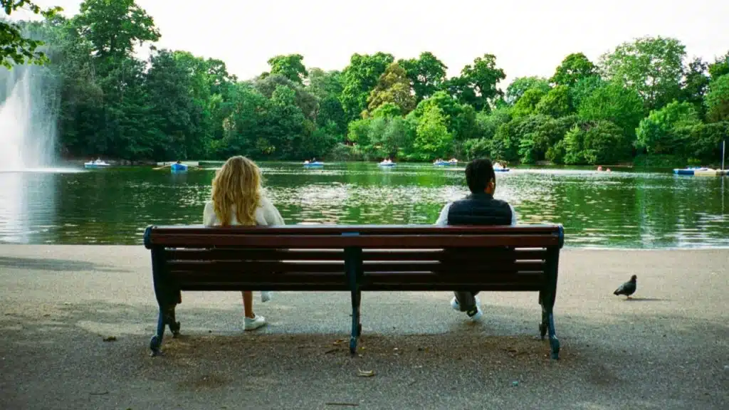A man and woman sit on a park bench facing a pond with their backs turned.