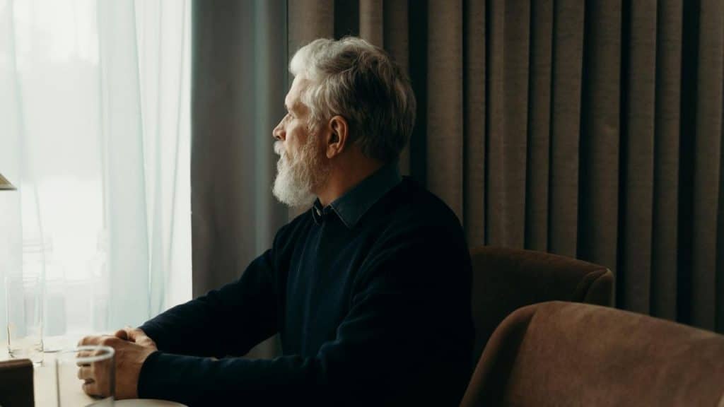 A man with grey hair and a beard sits at a table, looking out a window.