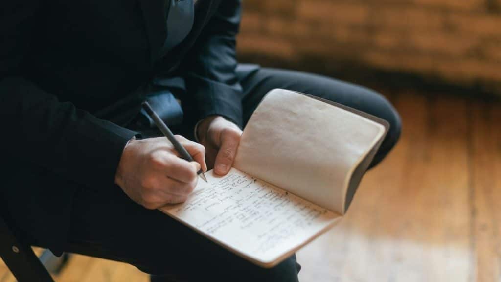 A person in a suit writes notes in a notebook while seated indoors.