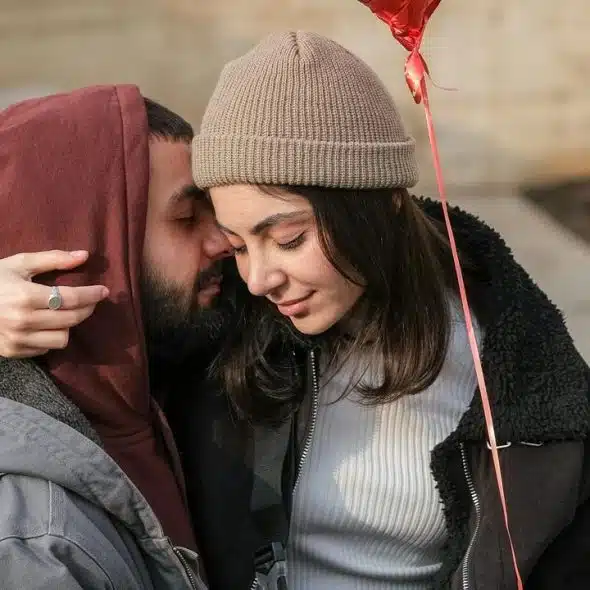 A couple cuddles closely outdoors, sharing a quiet moment while holding a red heart balloon.