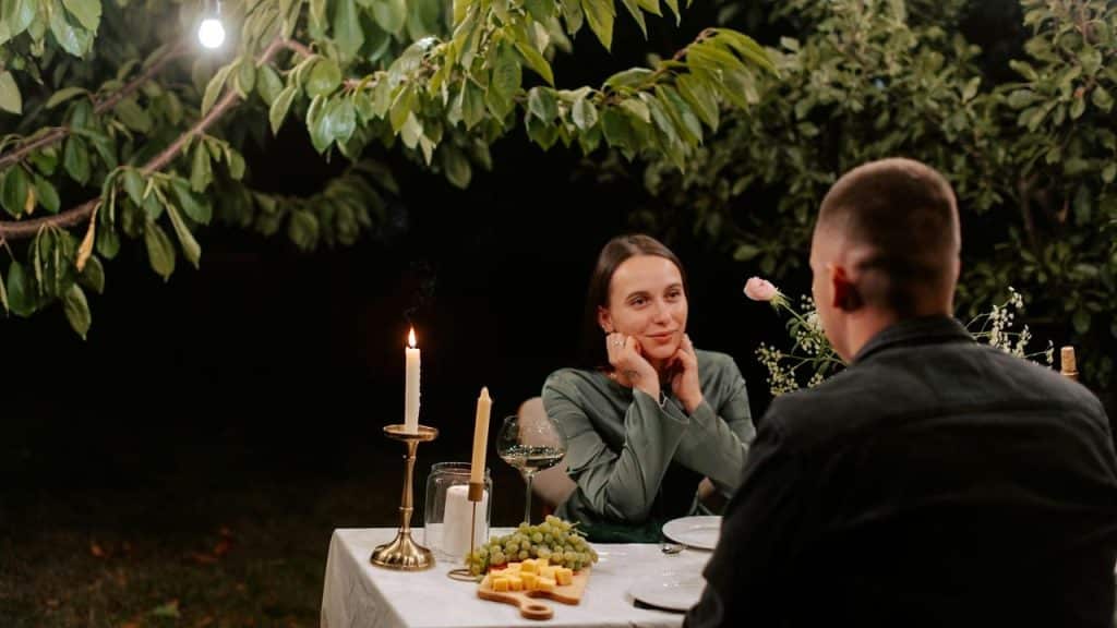 A couple enjoys a candlelit dinner outdoors under leafy trees at night.