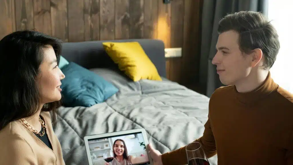 A couple video chats with a friend while holding wine glasses in a bedroom.