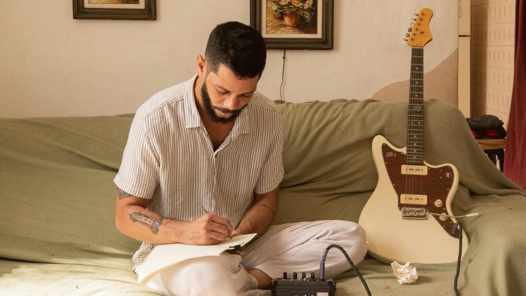 A man sits on a couch writing in a notebook beside an electric guitar.
