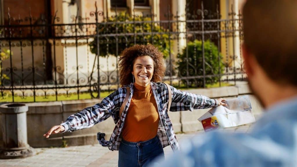 A smiling woman opens her arms while holding a map on a sunny city street.