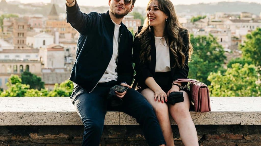 A couple laughs while taking a selfie with a city view in the background.