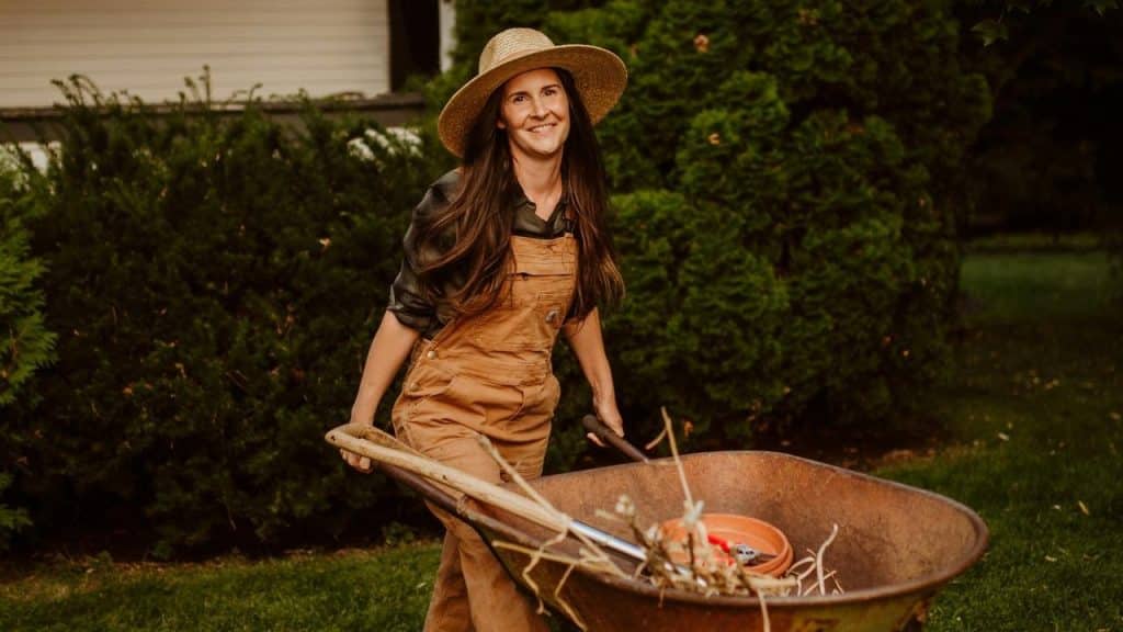 A smiling woman in overalls pushes a wheelbarrow in a garden.
