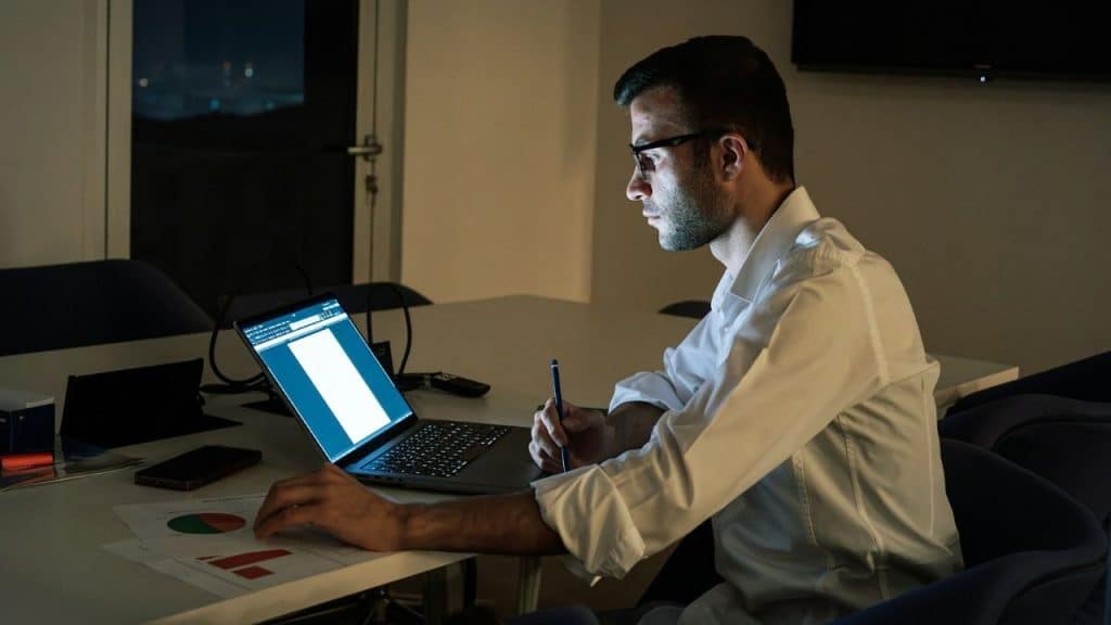 A man in a white shirt sits at a desk working on a laptop at night.