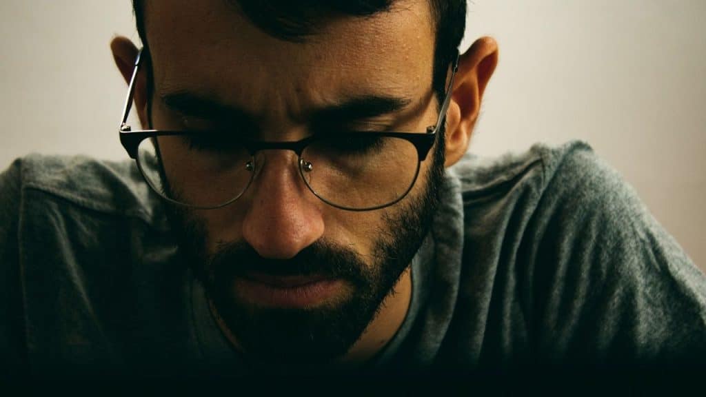 A close-up of a man wearing glasses with a beard looking down with a serious expression.