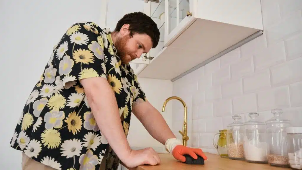 A man in a floral shirt leans over a wooden counter and wipes it with a sponge.