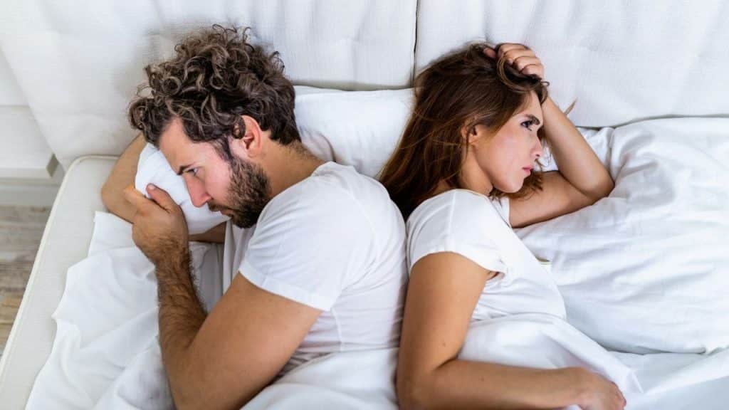 A man and a woman lie in bed facing away from each other with frustrated expressions.