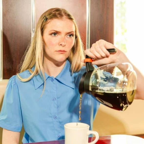 A woman with long blonde hair pours dark liquid from a glass pot into a mug.