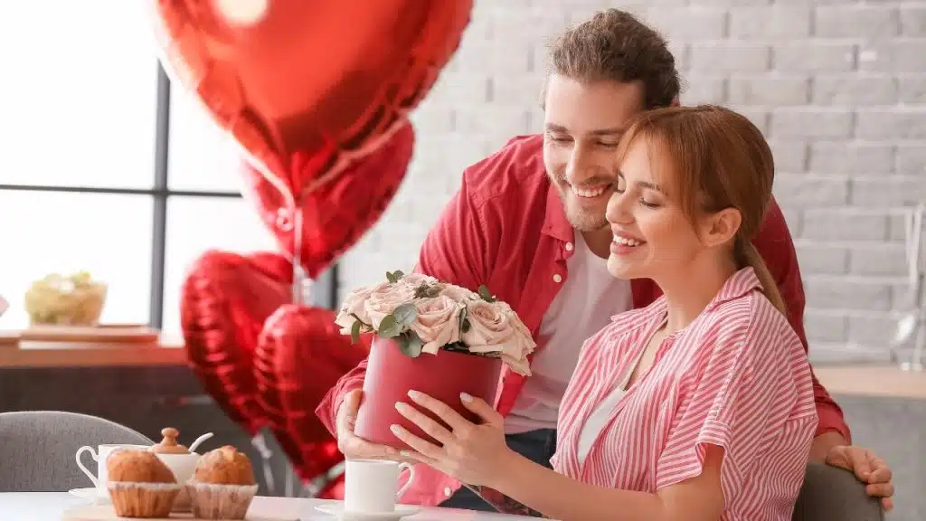 A man in a red shirt presents a round box of pink roses to a woman.