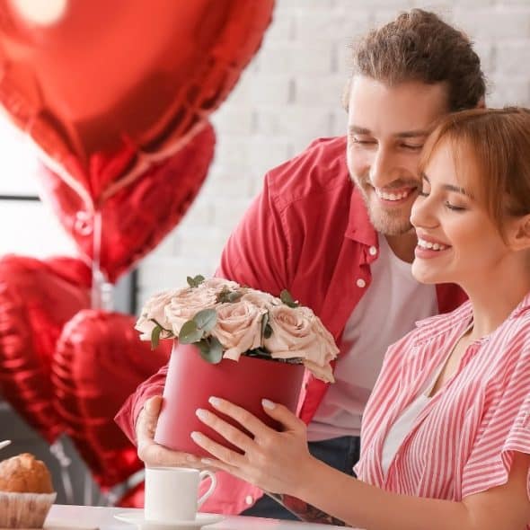 A man in a red shirt presents a round box of pink roses to a woman.