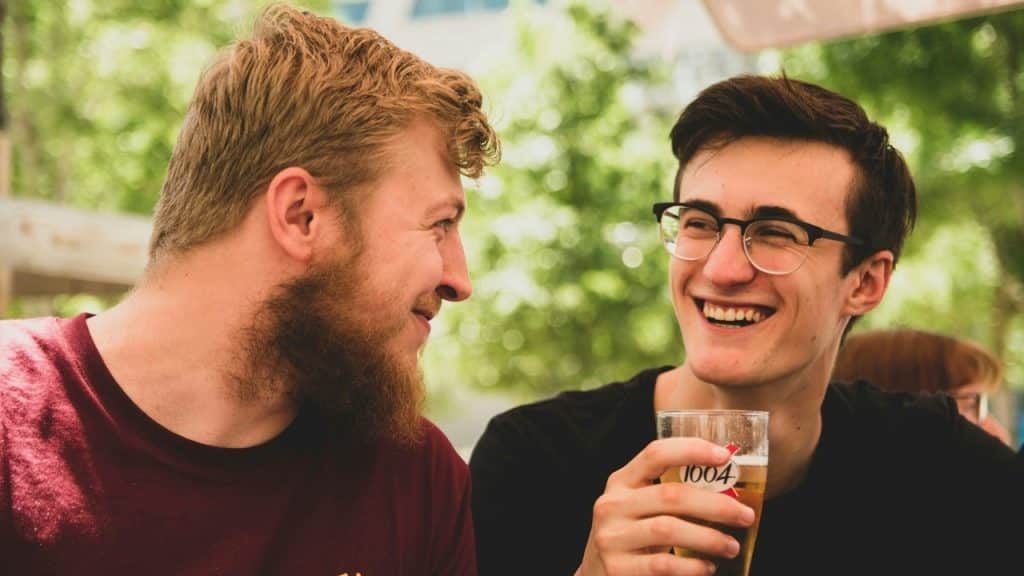 Two men sit outside, laughing together while one holds a glass containing a light beverage.