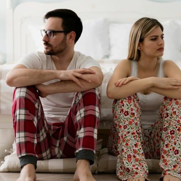 A couple wearing pajamas sitting on the the floors with their heads turned away from each other.