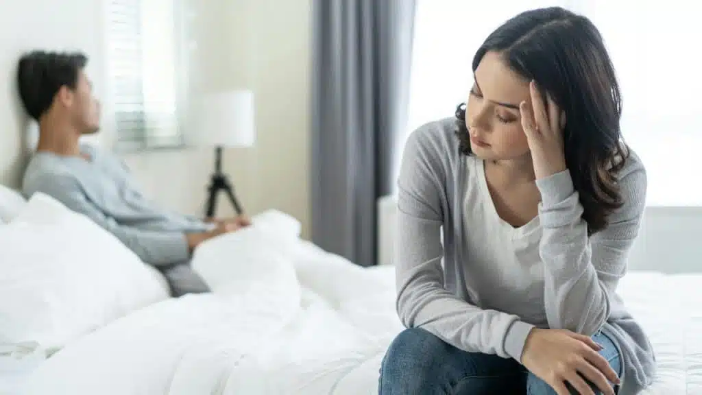 A woman sits on the edge of a bed with her hand on her forehead, looking distressed, while a man sits in the background facing away.