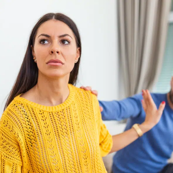 A woman in a yellow sweater looks away while gesturing for a man to stop.