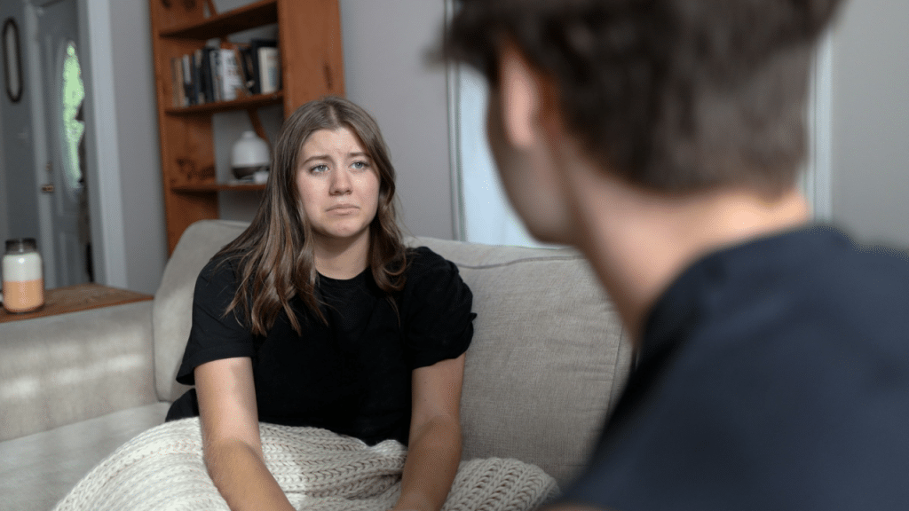 A woman with a sad expression sits on a couch while facing a blurry man.