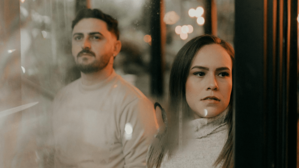 A man and woman stand behind a glass window looking out in different directions.
