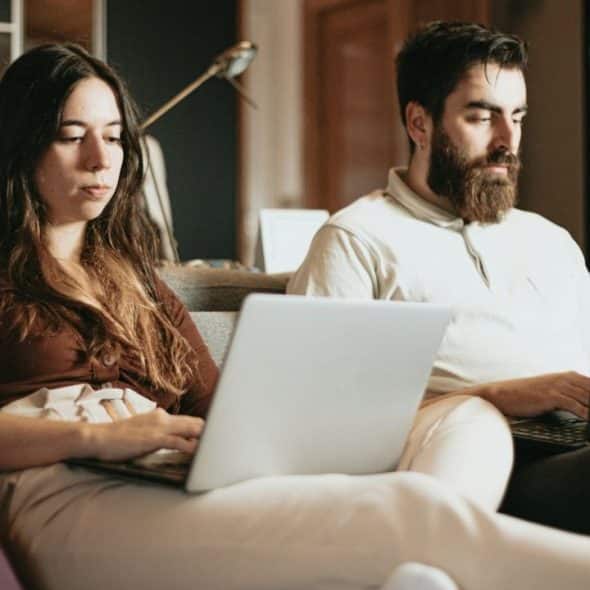 A bearded man and a woman with long hair sit on a couch using separate laptops.