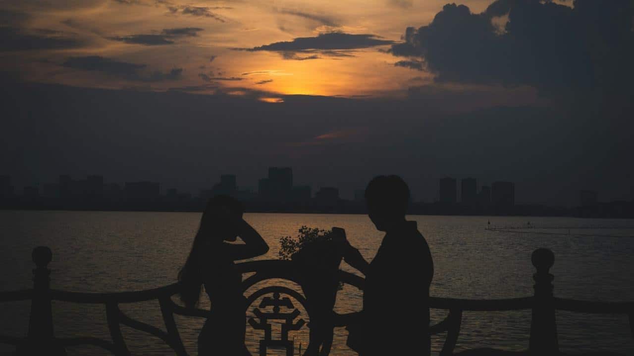 Silhouette of a couple standing by a decorative railing overlooking a body of water at sunset.